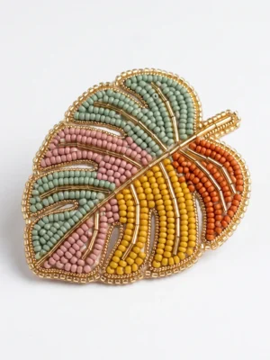 Hand beaded Monstera leaf brooch with pastel green, blush pink, mustard yellow and orange seed beads, finished with metallic gold bugle bead detailing.