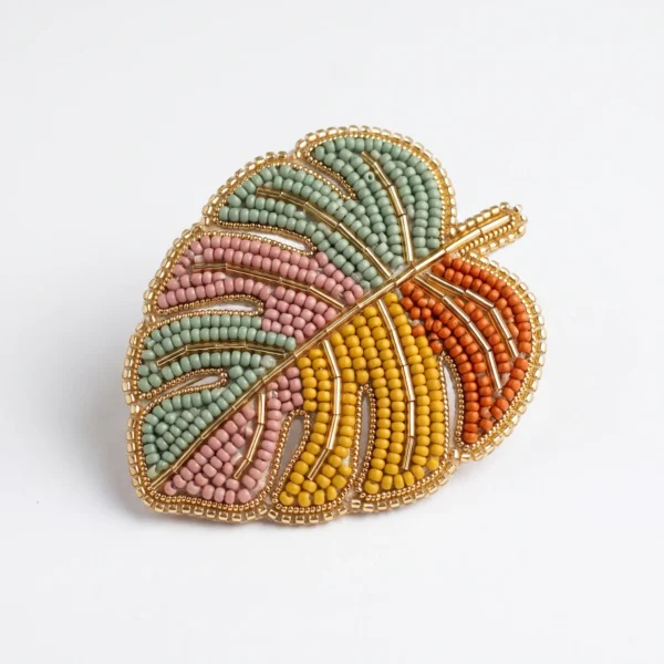 Hand beaded Monstera leaf brooch with pastel green, blush pink, mustard yellow and orange seed beads, finished with metallic gold bugle bead detailing.